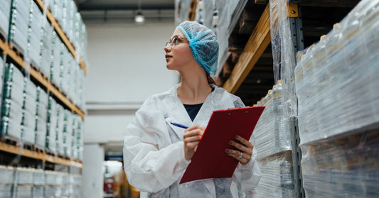 A woman in protective gear walking around a medical supplies warehouse. She is holding a clipboard in her hands.